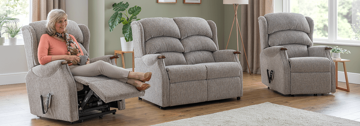 A person relaxing in a Celebrity Westbury recliner beside a matching sofa in a bright living room.